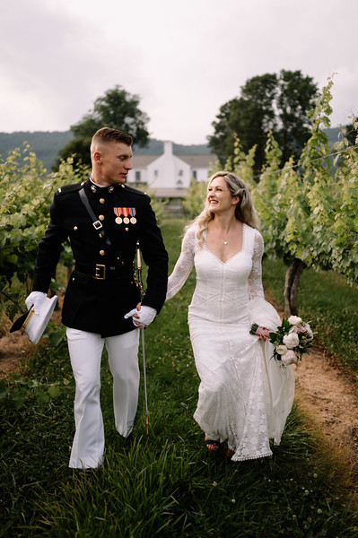 A bride in a white lace gown and a groom in a military dress uniform walking together in a vineyard.