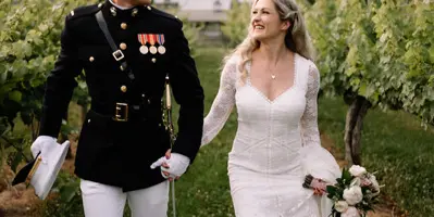 A bride in a white lace gown and a groom in a military dress uniform walking together in a vineyard.