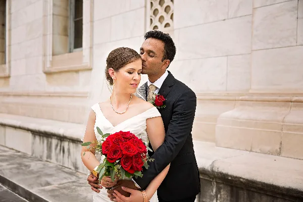 A bride in a white dress holding a bouquet of red roses is embraced by a groom in a black suit against a stone building facade.