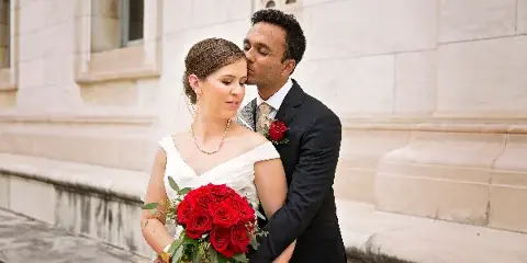 A bride in a white dress holding a bouquet of red roses is embraced by a groom in a black suit against a stone building facade.