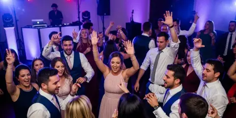 A crowded wedding dance floor with guests dancing and a DJ booth in the background.