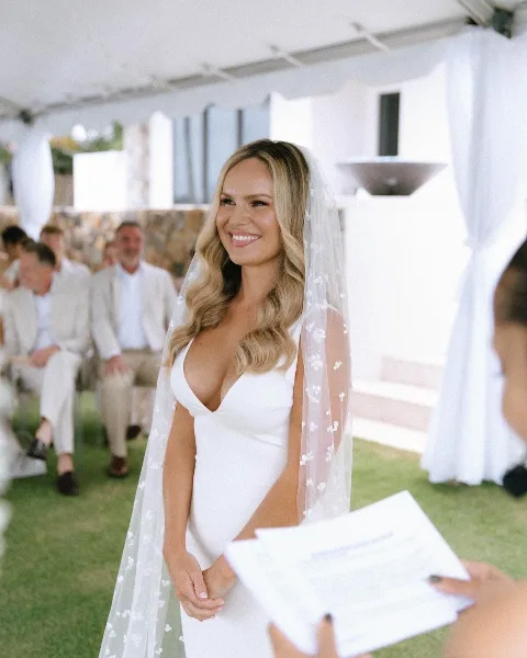 A smiling bride with a styled updo and professional makeup standing at an outdoor wedding ceremony.