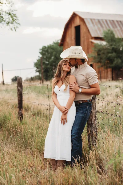 A couple posing for a portrait in a grassy field with a wooden barn in the background.
