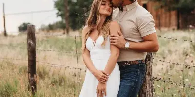 A couple posing for a portrait in a grassy field with a wooden barn in the background.