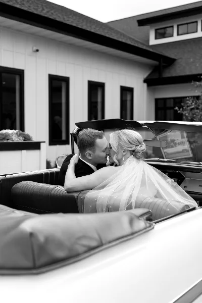 A black-and-white photograph of a bride and groom kissing inside a classic convertible car on their wedding day.