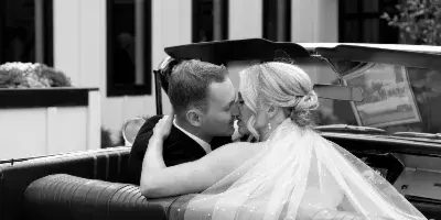 A black-and-white photograph of a bride and groom kissing inside a classic convertible car on their wedding day.