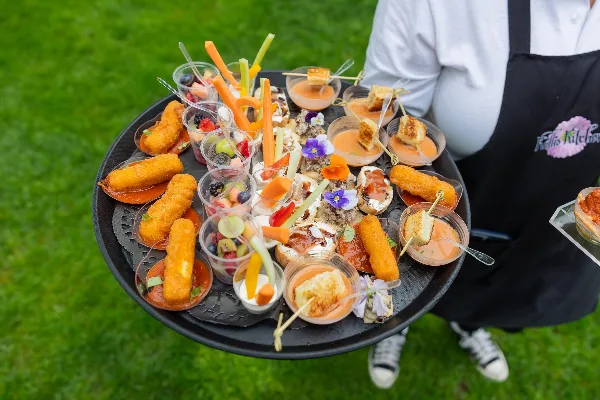 A catering staff member in a Kelli's Kitchen apron holding a tray of assorted appetizers and fruit cups outdoors.
