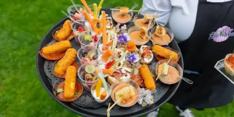 A catering staff member in a Kelli's Kitchen apron holding a tray of assorted appetizers and fruit cups outdoors.