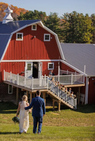 A bride and groom walking on a lawn toward a large red barn with white trim and a metal roof.