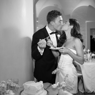 A black and white photograph of a bride and groom sharing a kiss while holding a cake and fork.