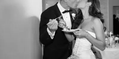 A black and white photograph of a bride and groom sharing a kiss while holding a cake and fork.