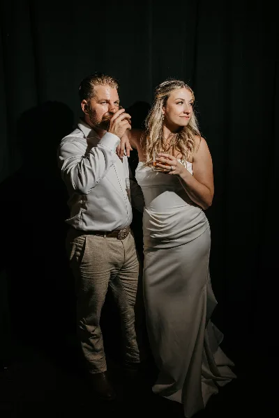 A moody, editorial-style photograph of a couple standing together against a dark background, with the woman wearing a white gown and holding a drink.
