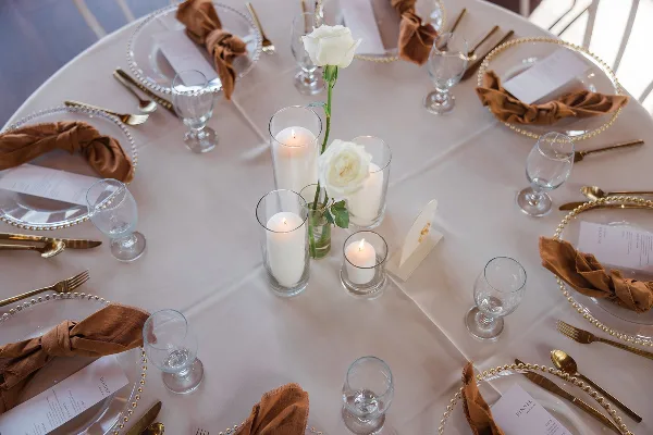 A close-up view of a formal table setting with white linens, gold-rimmed acrylic chargers, brown cloth napkins, and glass candle holders.