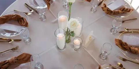 A close-up view of a formal table setting with white linens, gold-rimmed acrylic chargers, brown cloth napkins, and glass candle holders.