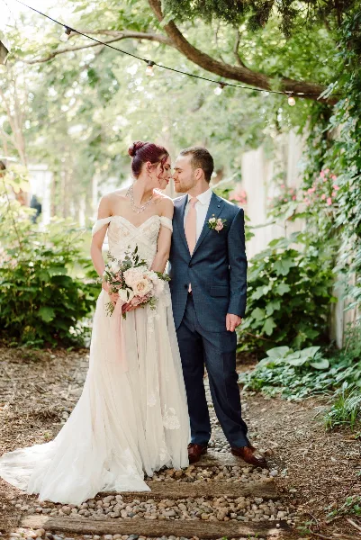A bride and groom standing close together in a garden, with the bride holding a bouquet of pink and white flowers.