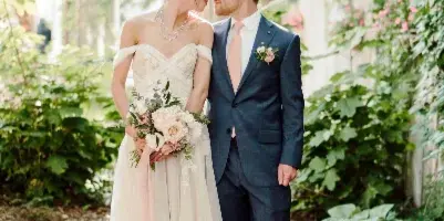 A bride and groom standing close together in a garden, with the bride holding a bouquet of pink and white flowers.