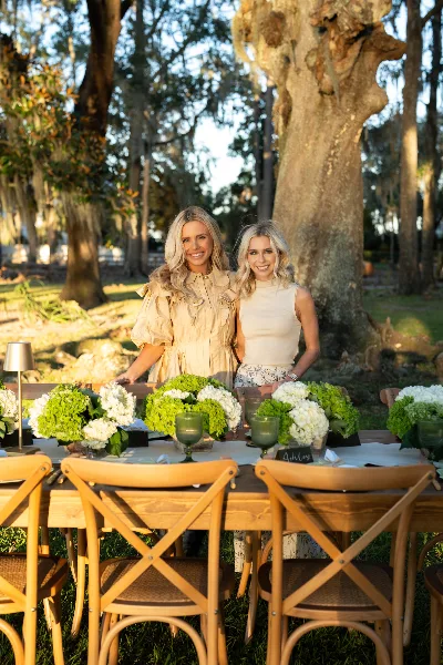 Two women standing behind an outdoor event table set with wooden chairs, a lamp, and floral centerpieces.