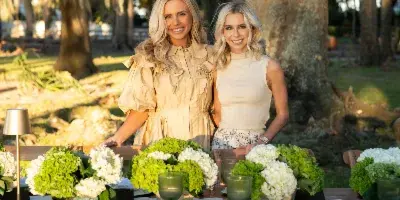 Two women standing behind an outdoor event table set with wooden chairs, a lamp, and floral centerpieces.