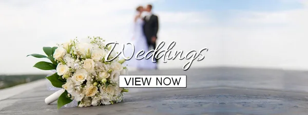 A white rose and lily wedding bouquet resting on a wooden surface with a blurred bride and groom in the background.