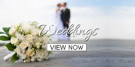 A white rose and lily wedding bouquet resting on a wooden surface with a blurred bride and groom in the background.