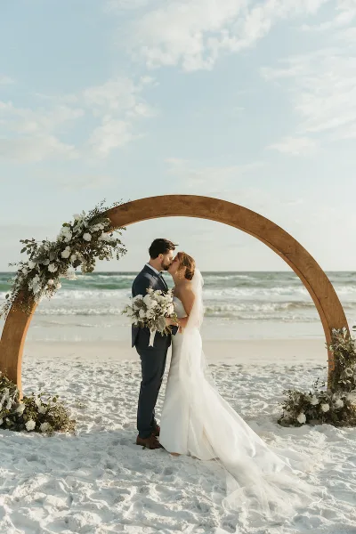A bride and groom kissing under a floral arch on a beach.