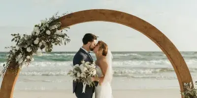 A bride and groom kissing under a floral arch on a beach.