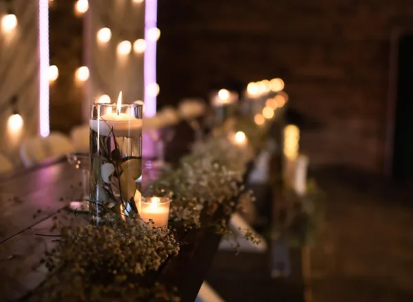 A close-up of a wedding table centerpiece featuring a tall glass candle holder, baby's breath, and string lights in a dimly lit venue.
