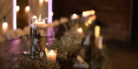 A close-up of a wedding table centerpiece featuring a tall glass candle holder, baby's breath, and string lights in a dimly lit venue.