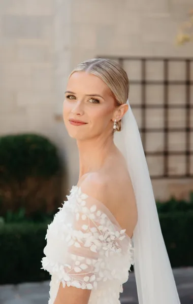 A bride with an elegant updo hairstyle and natural makeup, wearing a white floral dress and veil.