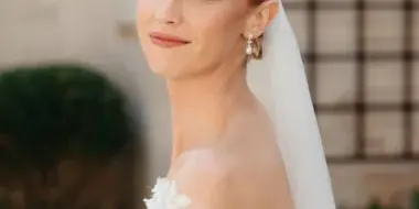 A bride with an elegant updo hairstyle and natural makeup, wearing a white floral dress and veil.