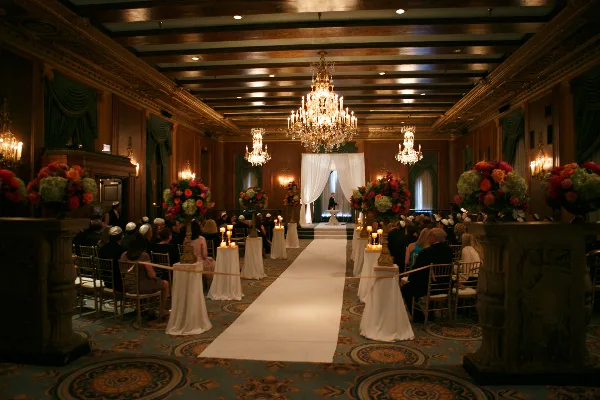 A formal wedding ceremony in progress inside an ornate ballroom with chandeliers and floral decorations.