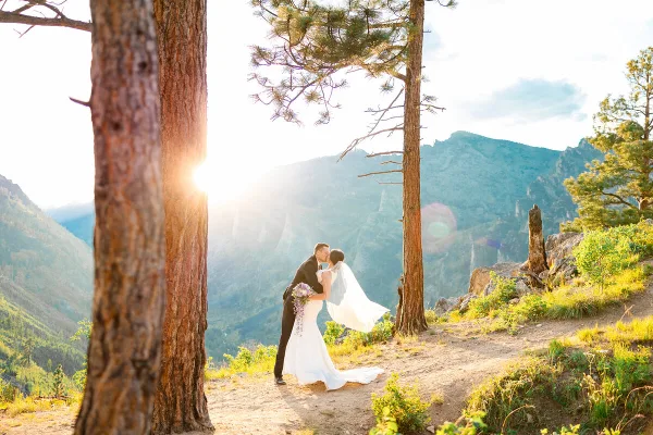 A bride and groom kissing on a mountain overlook with pine trees and a sunset.