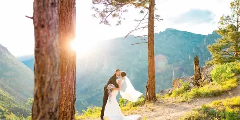 A bride and groom kissing on a mountain overlook with pine trees and a sunset.