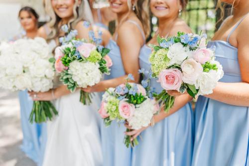 A bride and bridesmaids in blue dresses holding white, pink, and blue wedding bouquets.