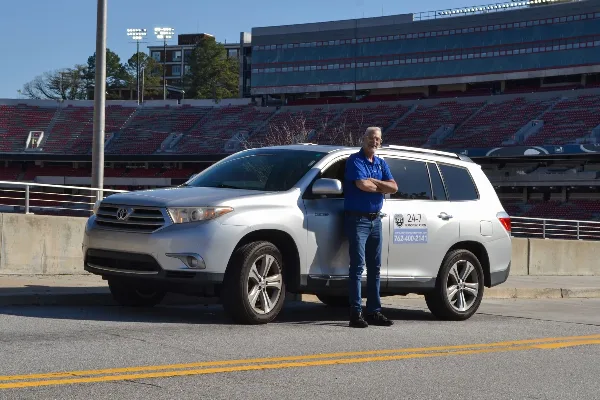 A man in a blue shirt stands with arms crossed next to a white Toyota SUV with Andy's Transport Services branding on the door, parked in front of a stadium.