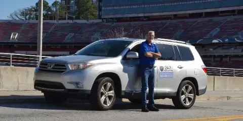 A man in a blue shirt stands with arms crossed next to a white Toyota SUV with Andy's Transport Services branding on the door, parked in front of a stadium.