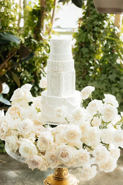A multi-tiered white wedding cake with detailed fondant work and sugar flowers, displayed on a gold stand surrounded by white roses and carnations.