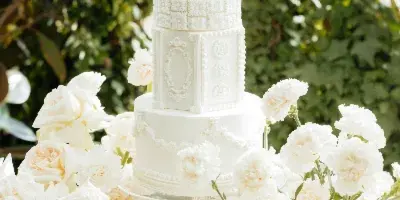 A multi-tiered white wedding cake with detailed fondant work and sugar flowers, displayed on a gold stand surrounded by white roses and carnations.