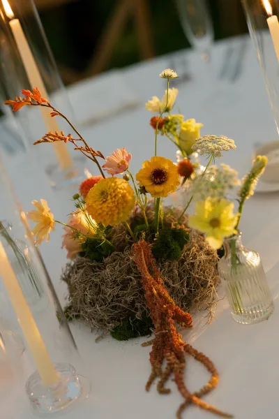 A close-up of a creative floral centerpiece featuring yellow and pink blooms, moss, and dried textures on a white table.
