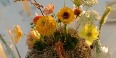A close-up of a creative floral centerpiece featuring yellow and pink blooms, moss, and dried textures on a white table.