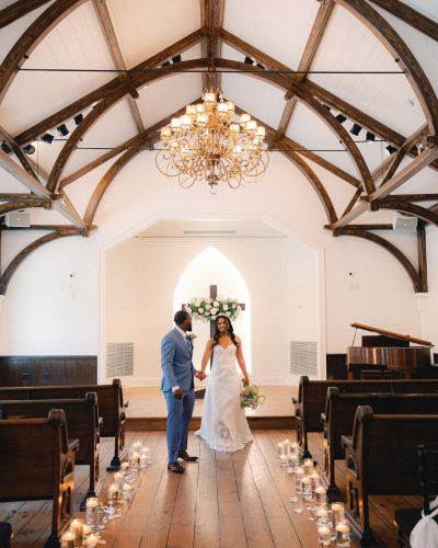 A bride and groom standing in the aisle of a chapel with a vaulted wooden ceiling and chandelier.