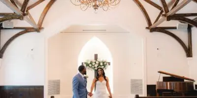 A bride and groom standing in the aisle of a chapel with a vaulted wooden ceiling and chandelier.