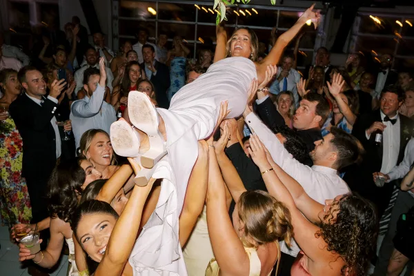 A bride in a white dress is lifted into the air by a crowd of cheering wedding guests.