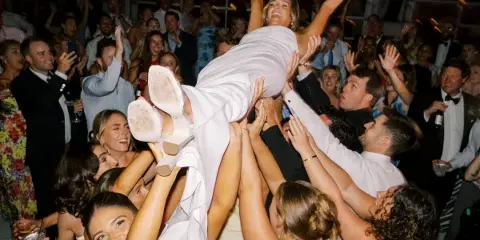 A bride in a white dress is lifted into the air by a crowd of cheering wedding guests.