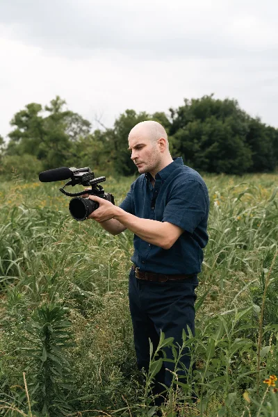 A bald man in a blue shirt holds a professional cinema camera with a microphone while standing in a field of tall grass.