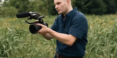 A bald man in a blue shirt holds a professional cinema camera with a microphone while standing in a field of tall grass.