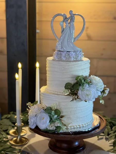 A two-tier white wedding cake decorated with white roses and a bride and groom topper, displayed on a wooden stand with lit candles.