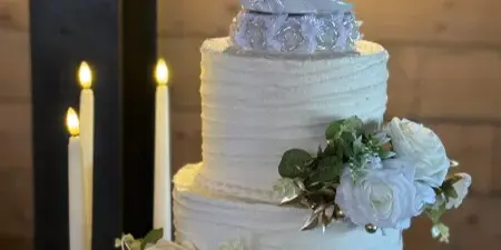 A two-tier white wedding cake decorated with white roses and a bride and groom topper, displayed on a wooden stand with lit candles.