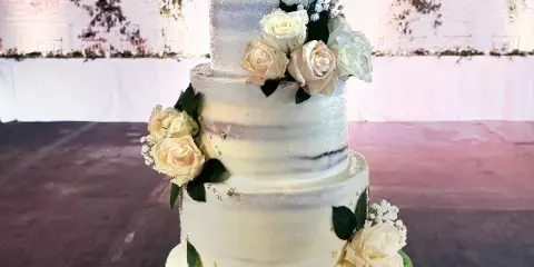 A three-tiered white wedding cake decorated with cream roses and greenery, set in a banquet hall.