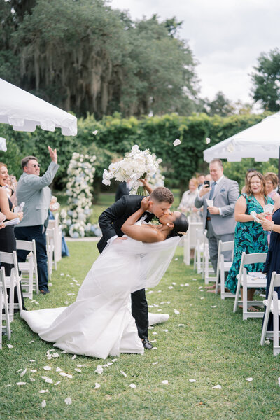 A bride and groom share a kiss during an outdoor wedding ceremony on a grassy lawn, surrounded by guests and white floral arches.